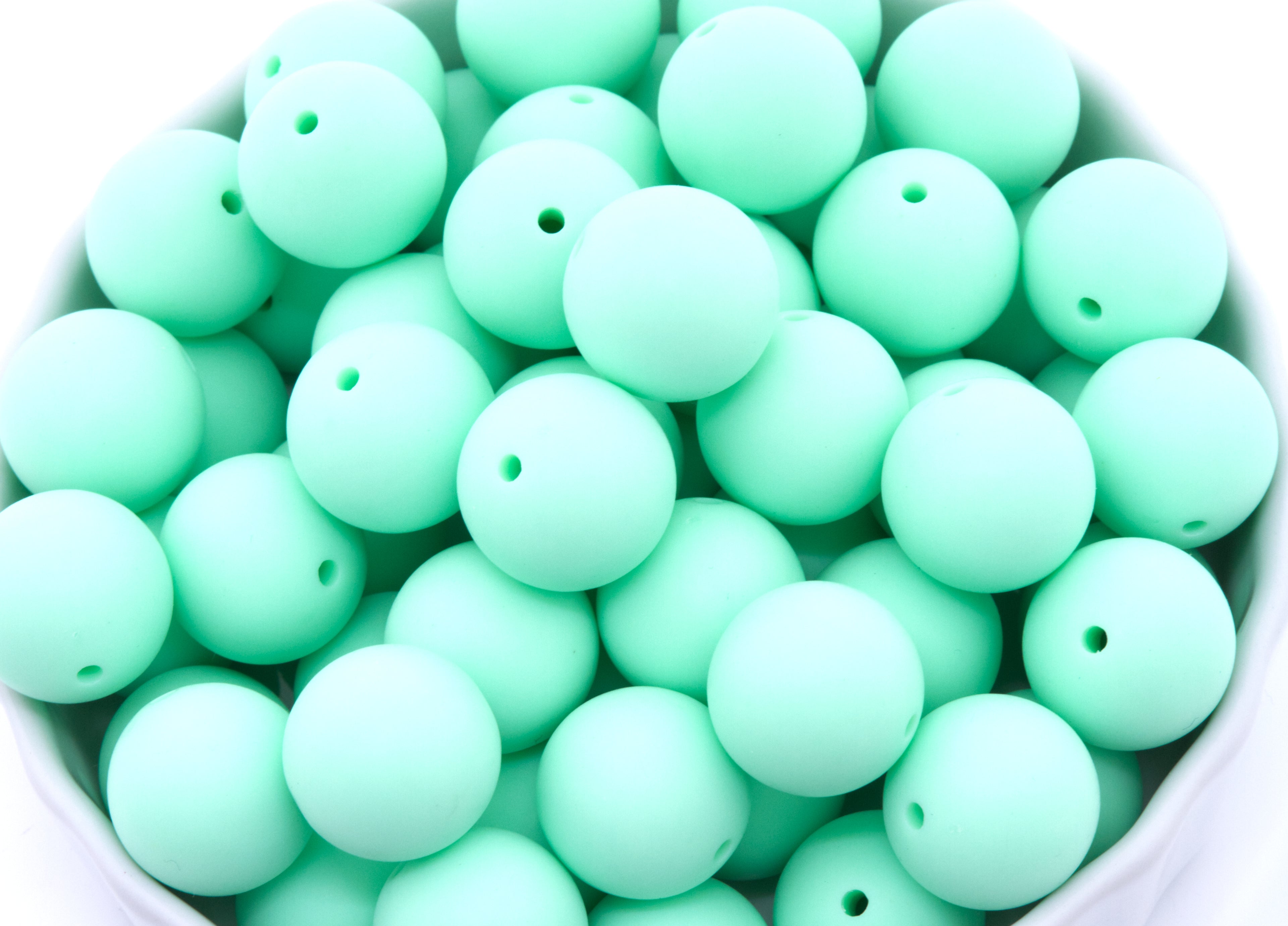 A close-up of many matte mint-green round beads, piled inside a white bowl against a white background.