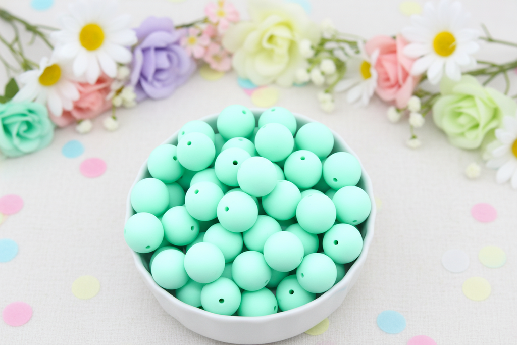 A white bowl filled with mint green round beads, set against a light background with pastel confetti and flowers.