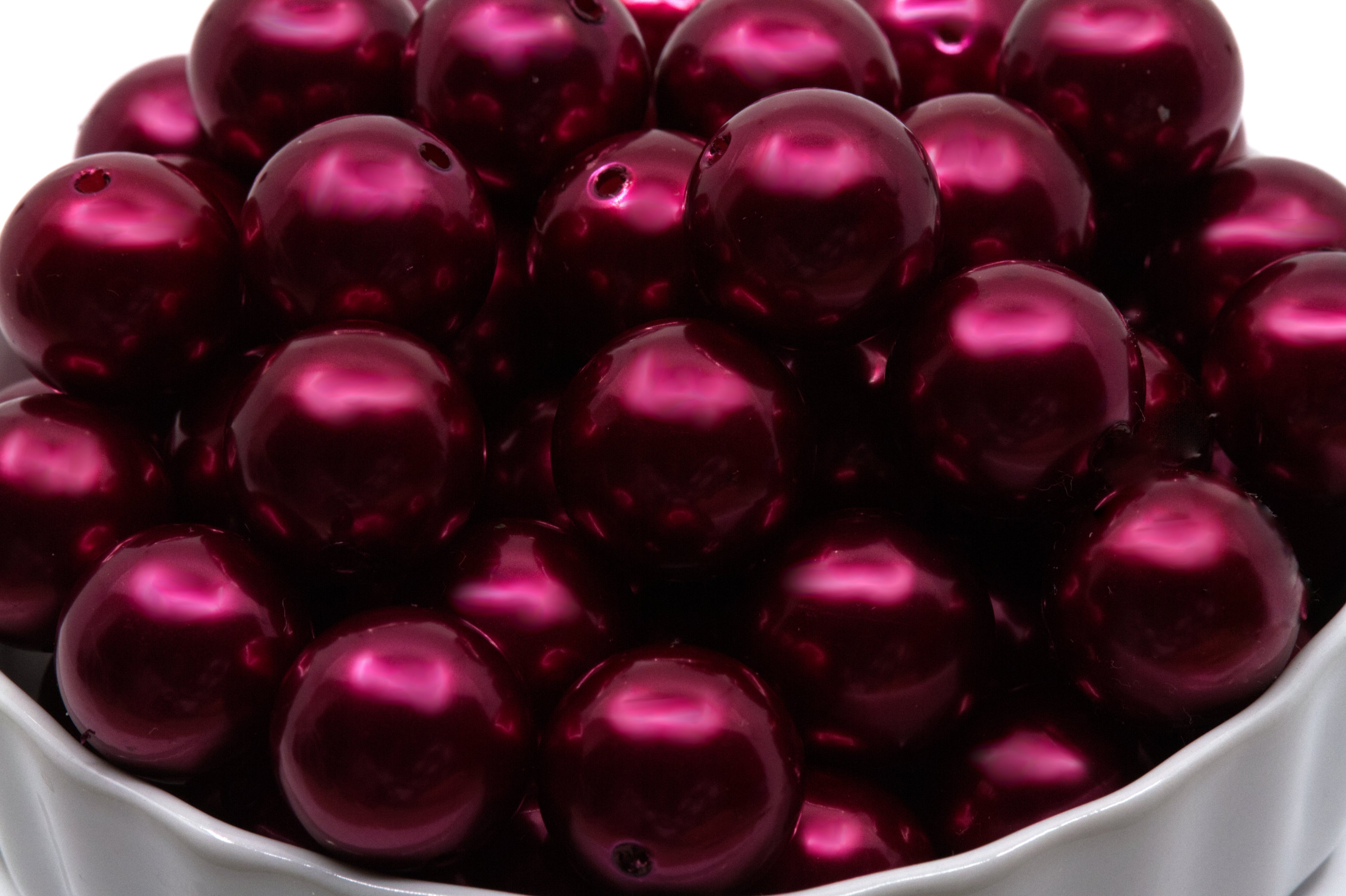 A close-up of glossy metallic deep red bubblegum beads piled in a white bowl against a white background.