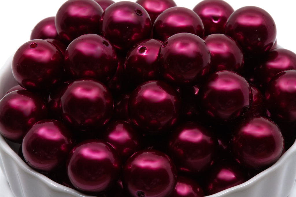 A close-up of glossy, metallic deep red bubblegum beads piled inside a white bowl against a white background.