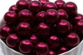 A close-up of glossy, metallic deep red bubblegum beads piled inside a white bowl against a white background.