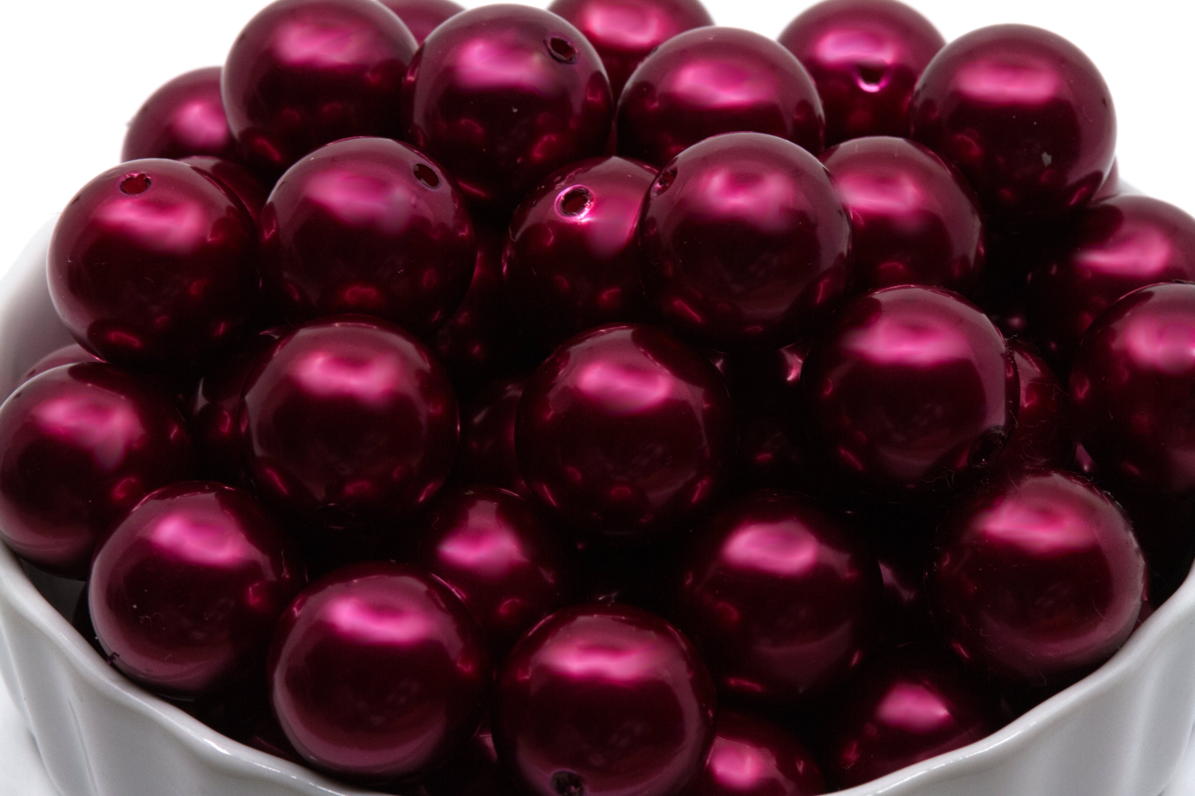 A close-up of glossy, metallic deep red bubblegum beads piled inside a white bowl against a white background.