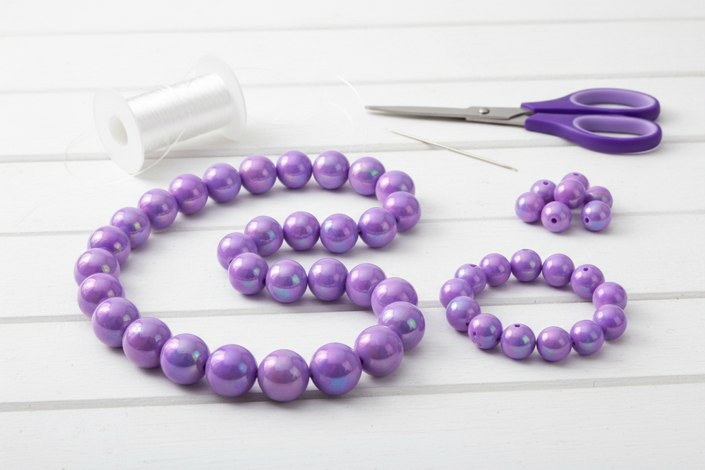 Purple iridescent beads, scissors, and a spool of thread on a white wooden surface