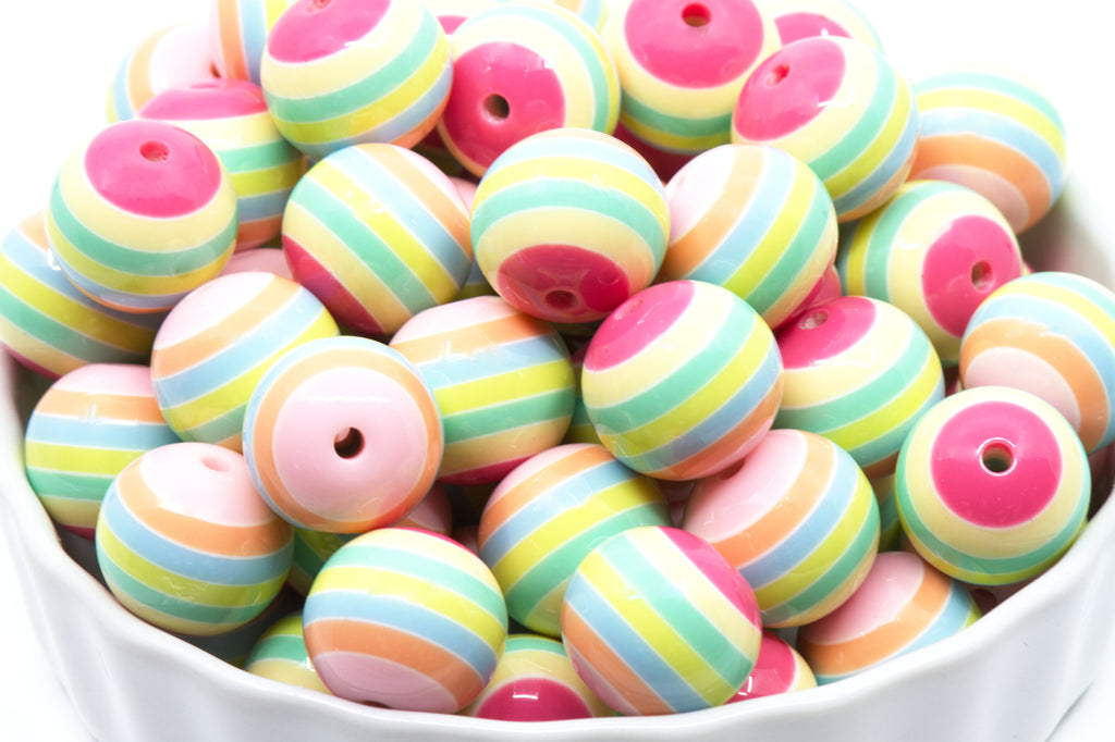 A pile of round, pastel-striped beads in a white bowl. The beads feature horizontal stripes of pink, light blue, mint green, yellow, and peach, and pink.