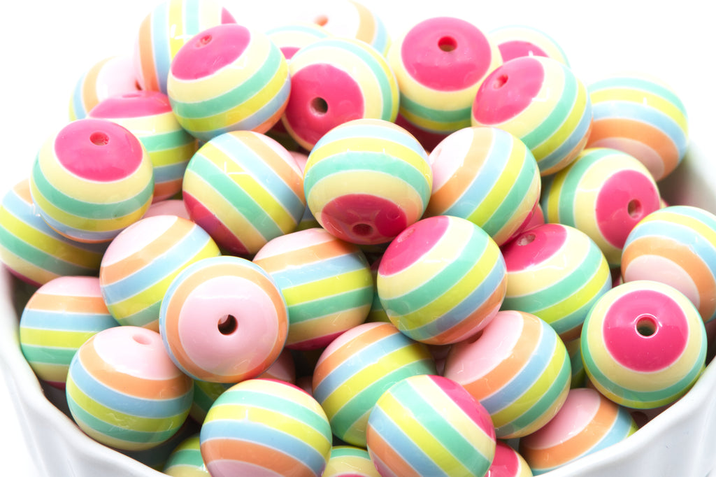 A pile of round, pastel-striped beads in a white bowl. The beads feature horizontal stripes of pink, light blue, mint green, yellow, and peach, and pink.
