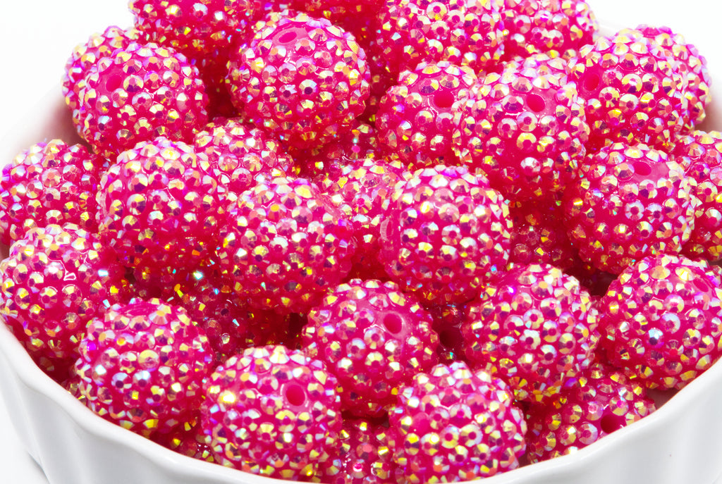 A close-up of bright pink rhinestone-covered round beads or embellishments piled in a white bowl against a white background.