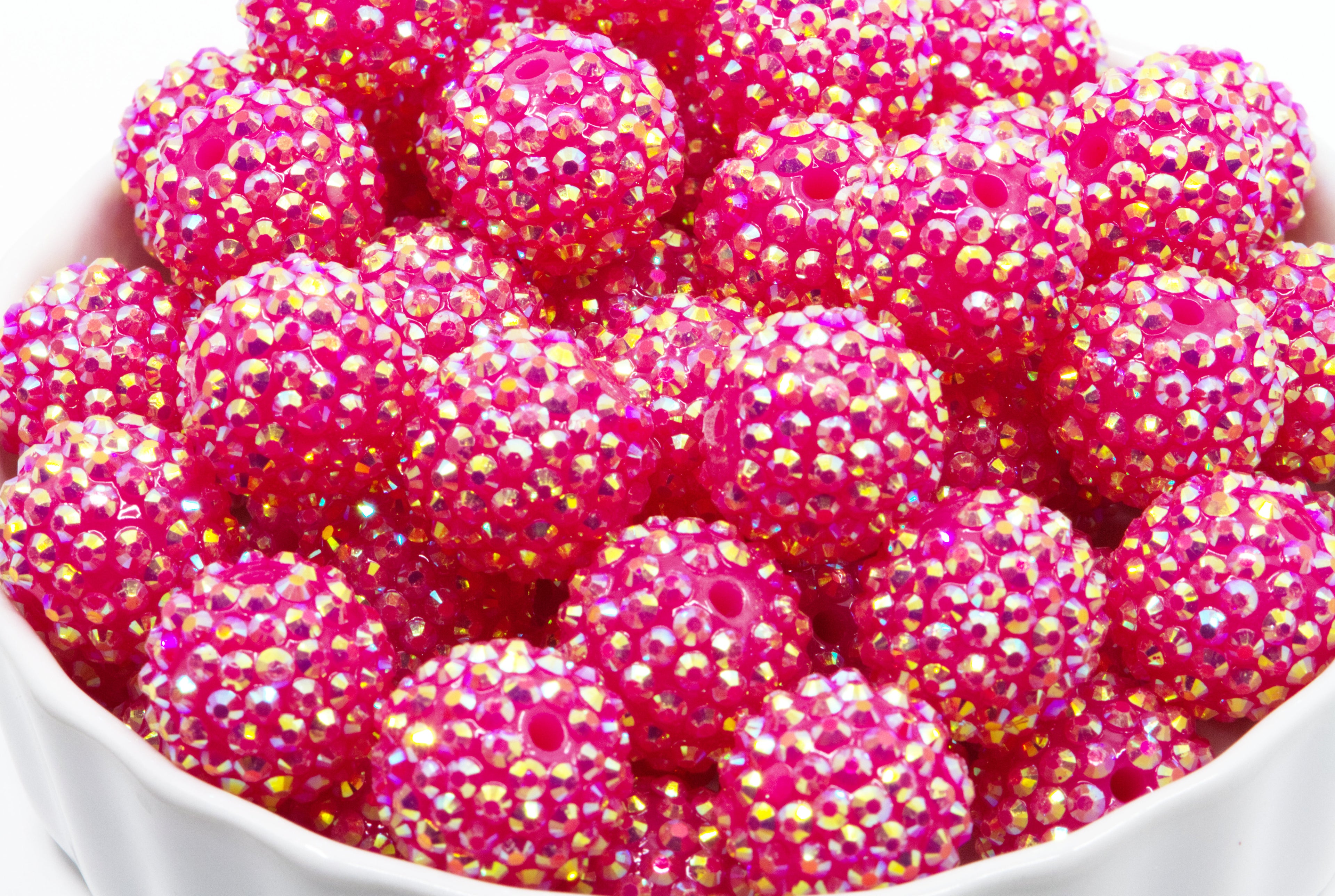 A close-up of bright pink rhinestone-covered round beads or embellishments piled in a white bowl against a white background.