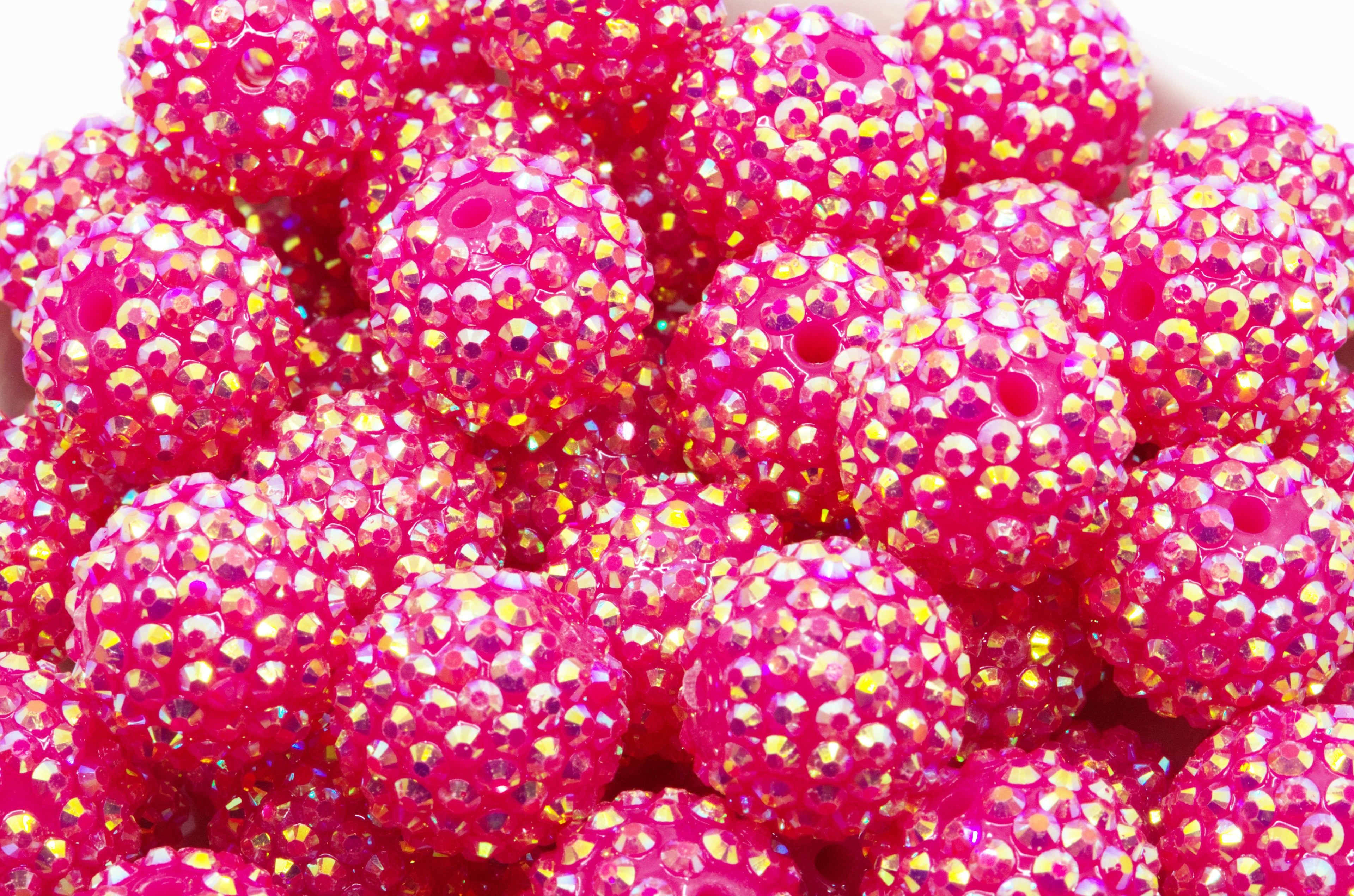 A close-up of bright pink rhinestone-covered round beads or embellishments piled in a white bowl against a white background.
