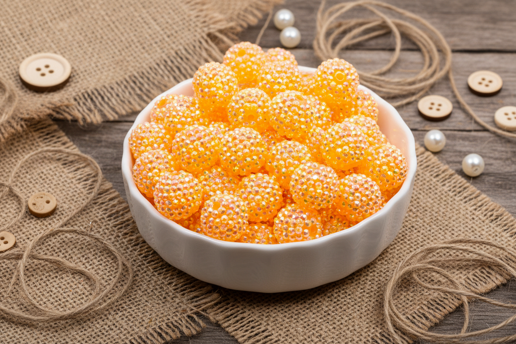 White bowl filled with orange rhinestone beads on a rustic wooden surface with buttons and twine.