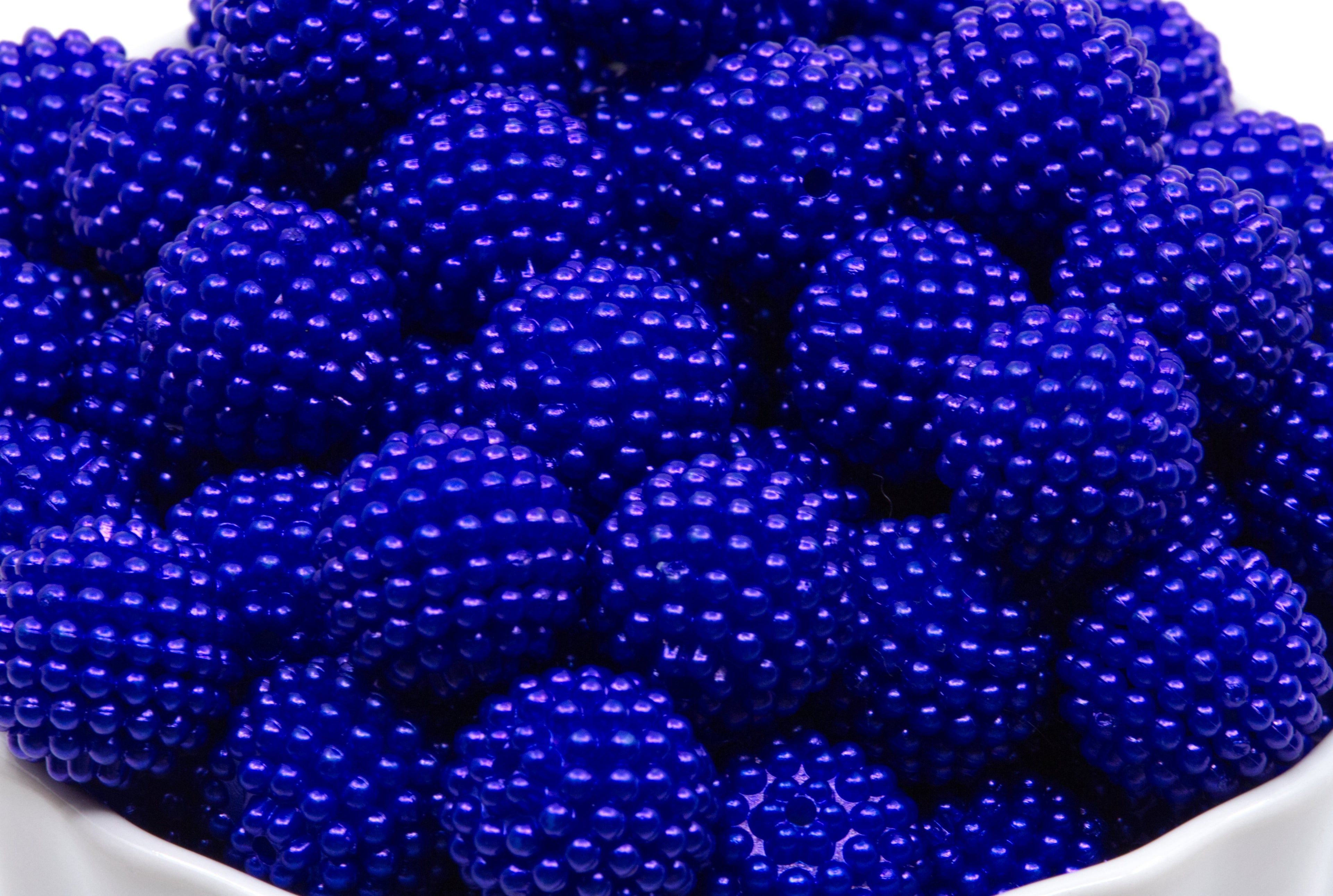 A close-up of blue beaded bubblegum beads piled in a white bowl against a white background.