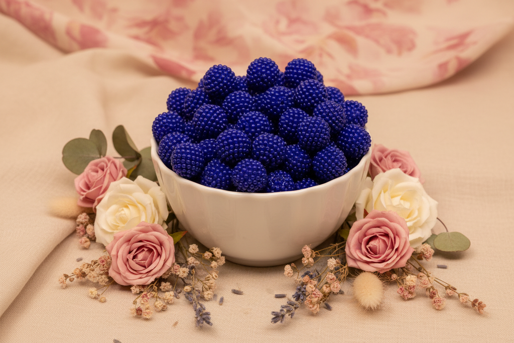 A white ceramic bowl filled with blue beaded bubblegum beads, styled with pink and white roses, dried flowers, and green leaves on a beige fabric surface.