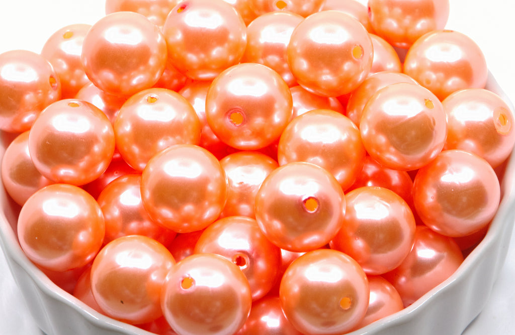 A close-up of many glossy sherbet orange shiny pearl beads piled in a white bowl against a white background.