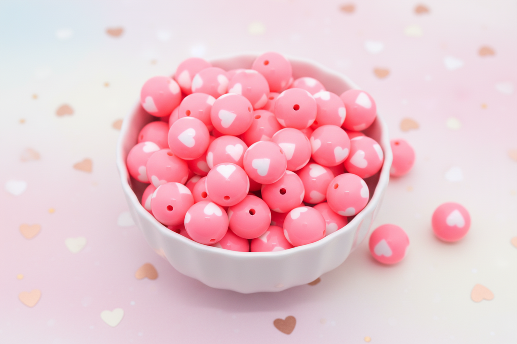 White bowl filled with pink beads with a heart pattern on a pink background