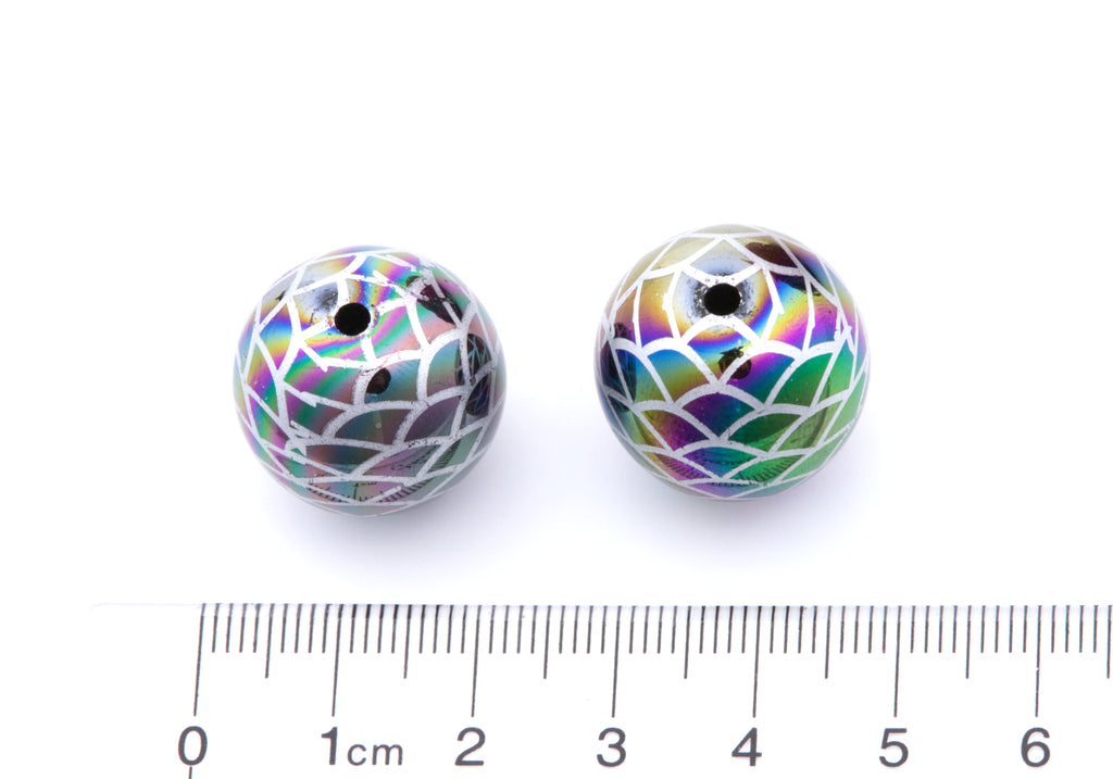 Two round iridescent beads with a mermaid scale pattern next to a cm ruler on a white background. 