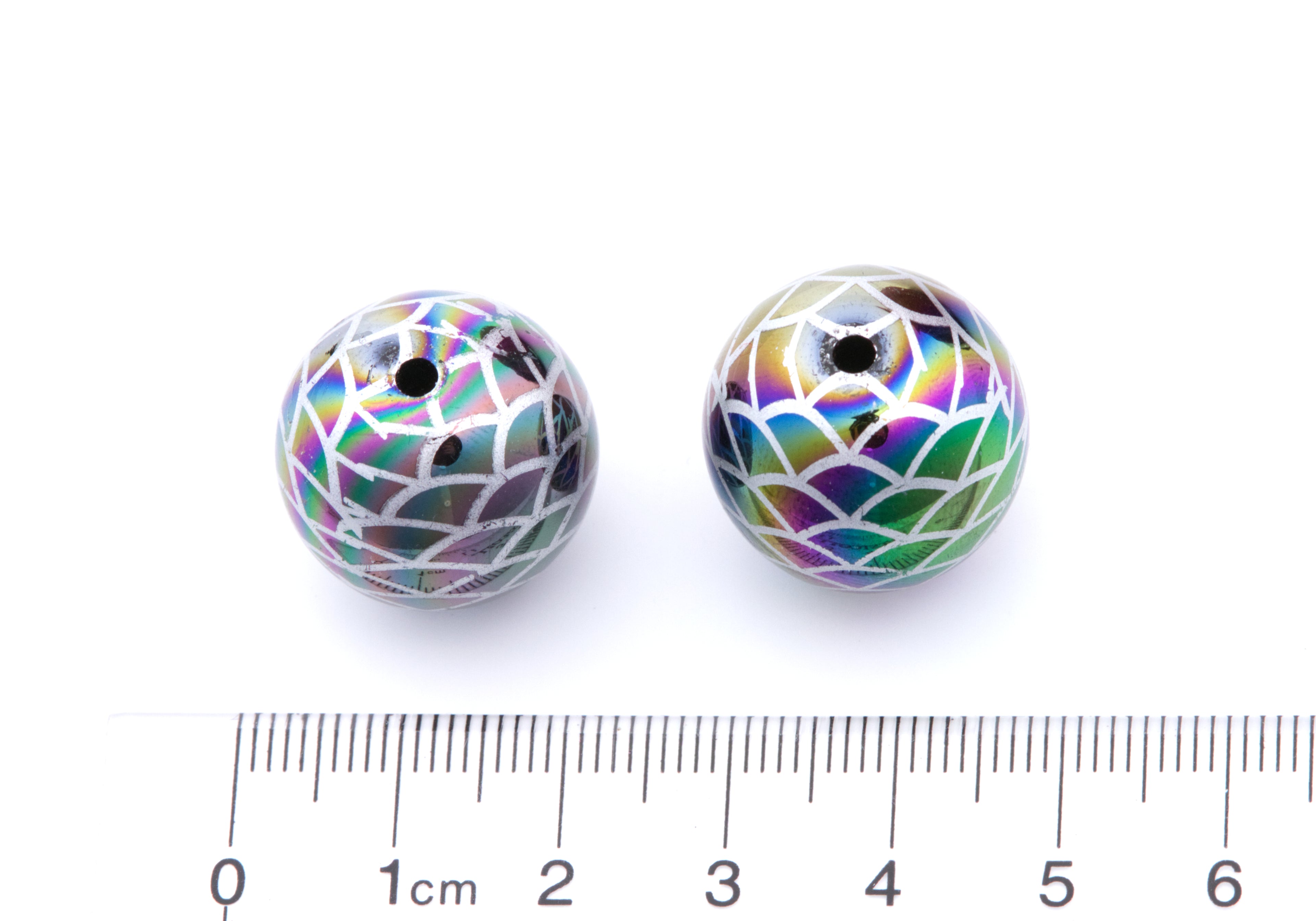 Two round iridescent beads with a mermaid scale pattern next to a cm ruler on a white background. 