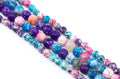 Multiple strands of round faceted agate beads in assorted colors including purple, blue, pink and white, arranged diagonally on a white background.