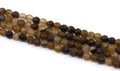 A close-up of multiple strands of faceted round agate beads in earthy brown, tan, and dark brown tones, arranged diagonally on a plain white background.