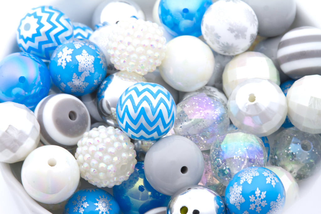 A close-up pile of decorative round beads in white, blue, gray, and iridescent finishes, including patterns like snowflakes, stripes, chevrons, and glitter.