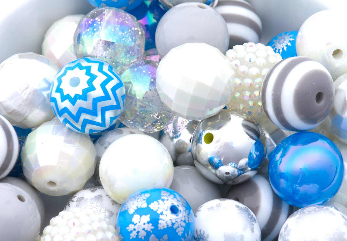 A close-up pile of decorative round beads in white, blue, gray, and iridescent finishes, including patterns like snowflakes, stripes, chevrons, and glitter.