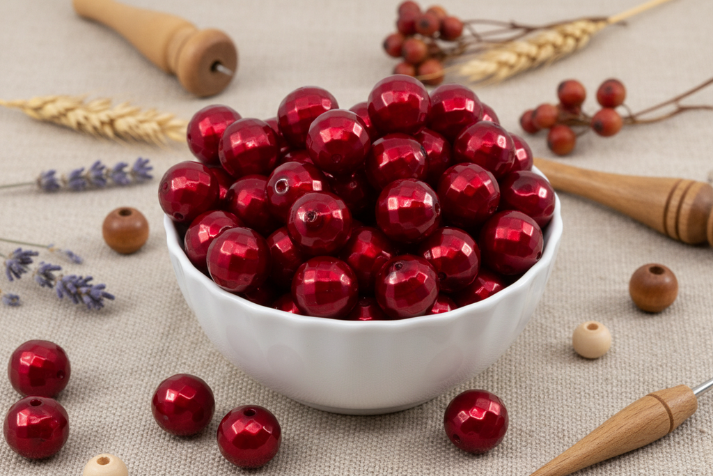 A white ceramic bowl filled with faceted red bubblegum beads, styled on a beige linen fabric with wheat stalks, lavender sprigs, and wooden tools.