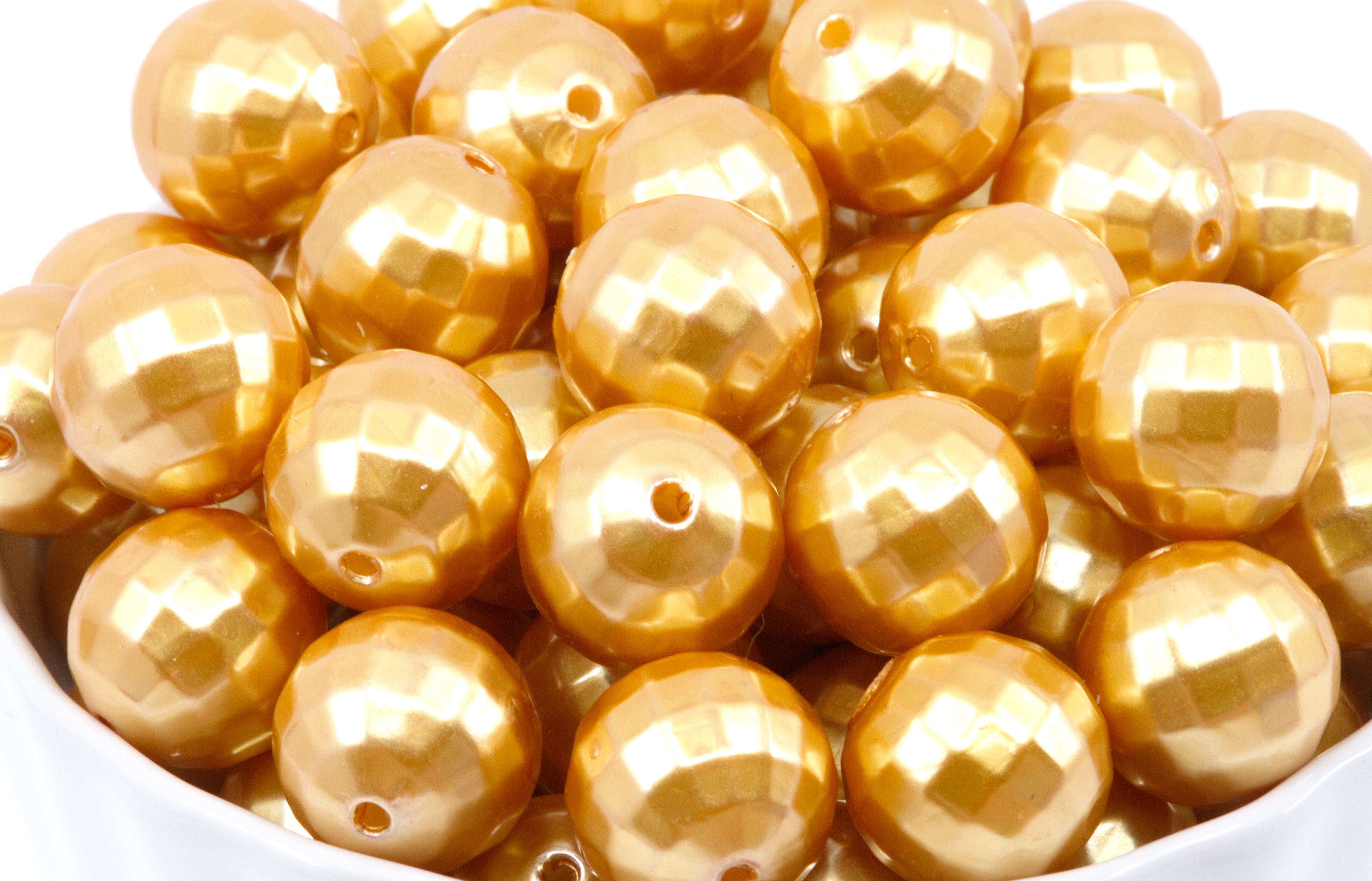 A pile of shiny metallic gold faceted round beads with holes, shown close up in a white container against a white background.