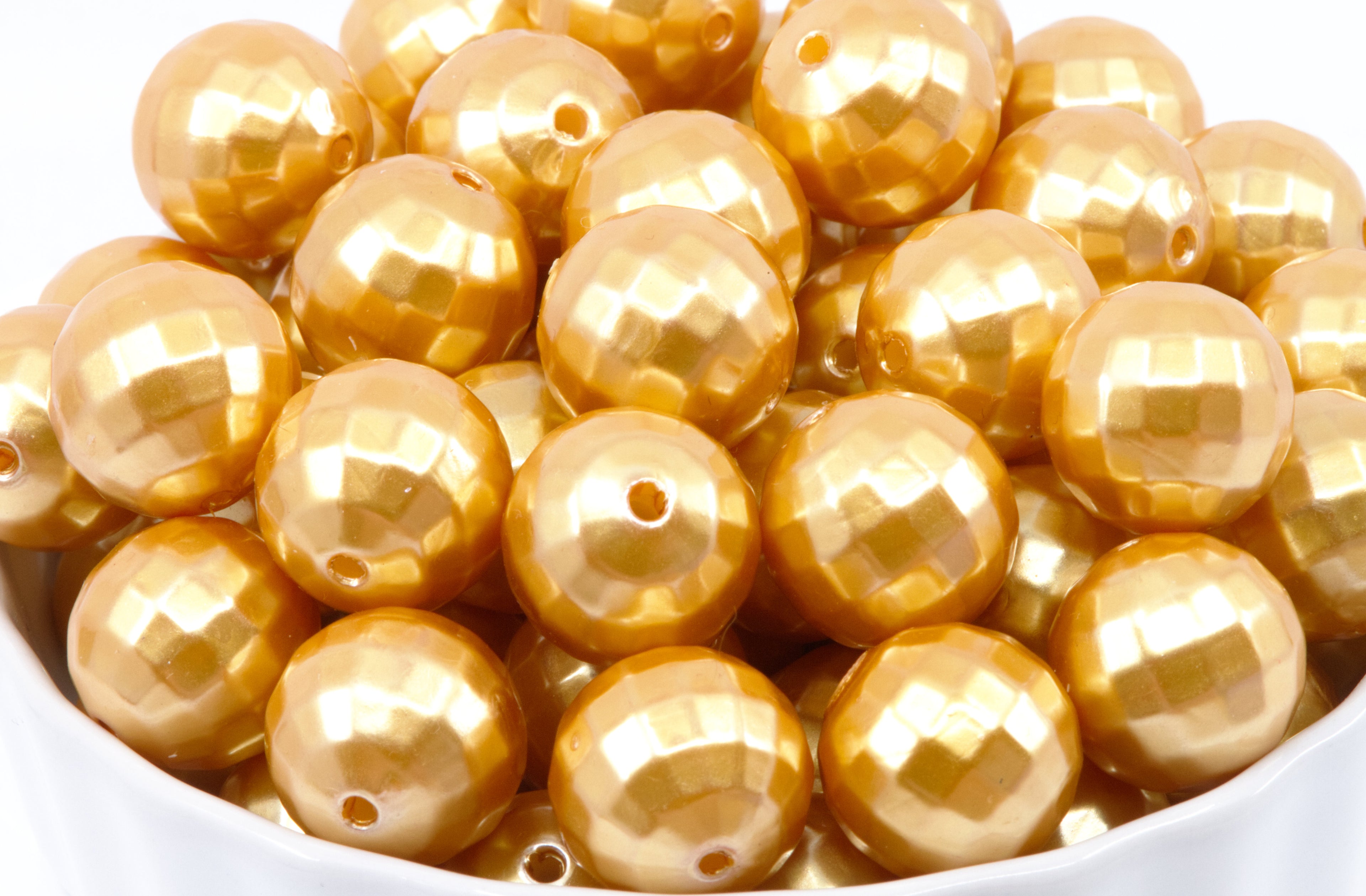 A pile of shiny metallic gold faceted round beads with holes, shown close up in a white container against a white background.