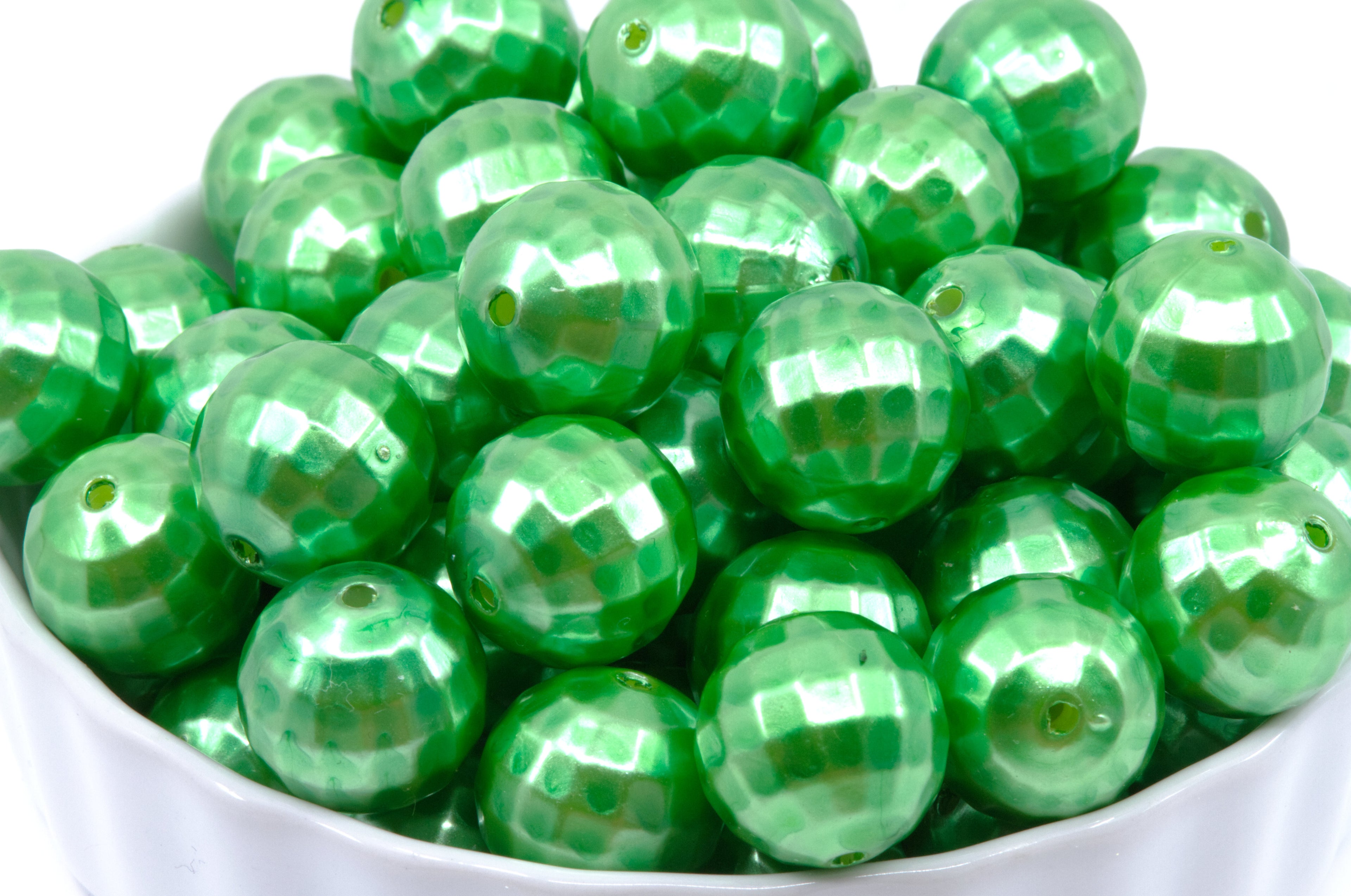 A close-up of shiny green faceted round beads piled in a white bowl against a white background.