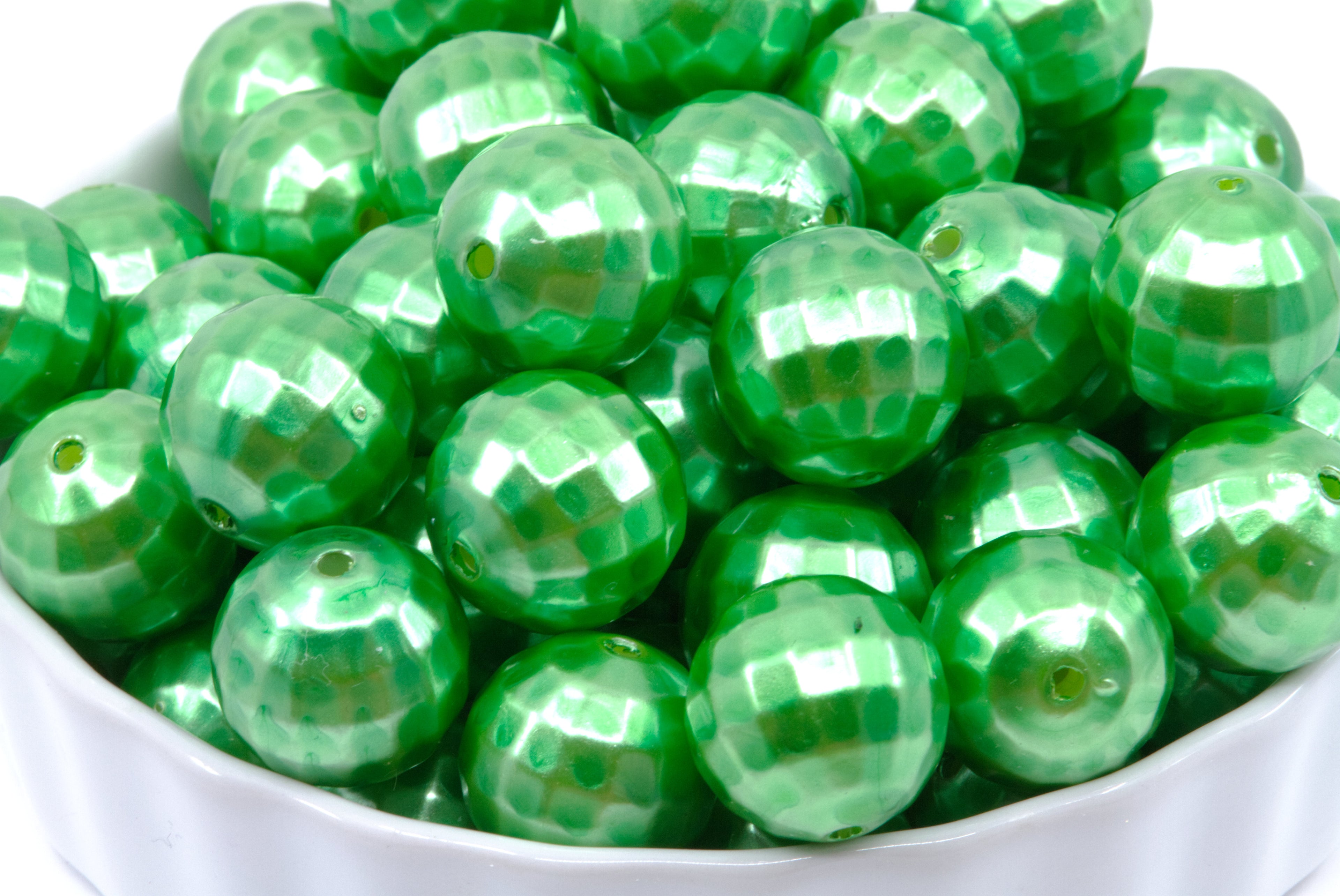 A close-up of shiny green faceted round beads piled in a white bowl against a white background.