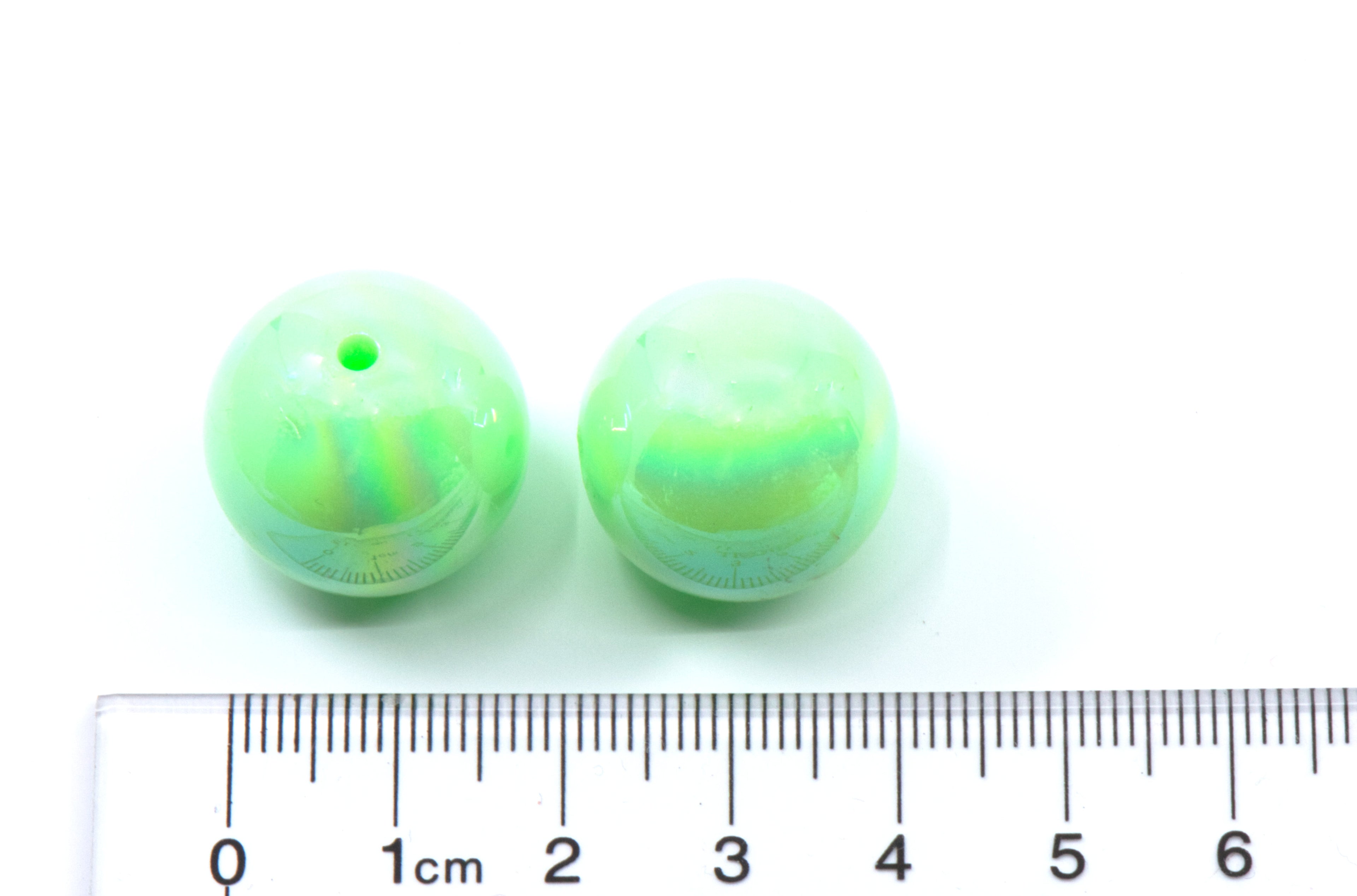 two green iridescent beads with a ruler for scale on a white background