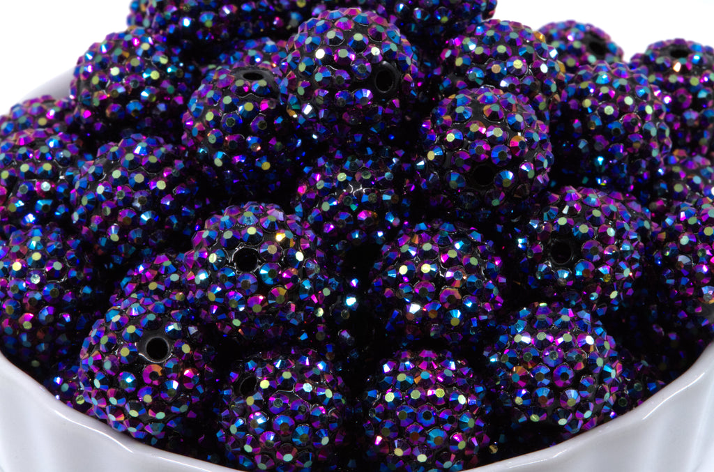 A close-up of round metallic indigo AB rhinestone bubblegum beads, piled inside a white bowl against a white background.