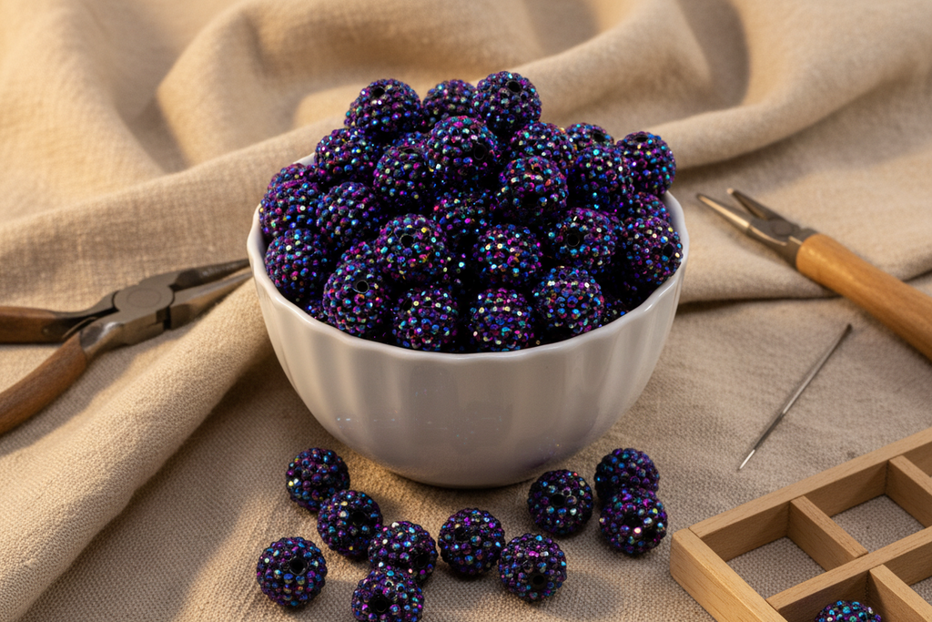 A white bowl filled with metallic indigo AB rhinestone bubblegum beads, styled on a beige linen cloth with crafting tools like pliers, a wooden needle, and a wooden bead grid.