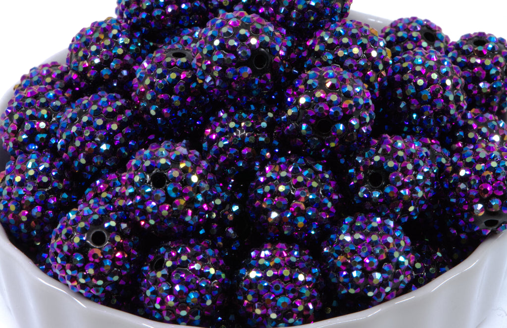 A close-up of round metallic indigo AB rhinestone bubblegum beads, piled inside a white bowl against a white background.