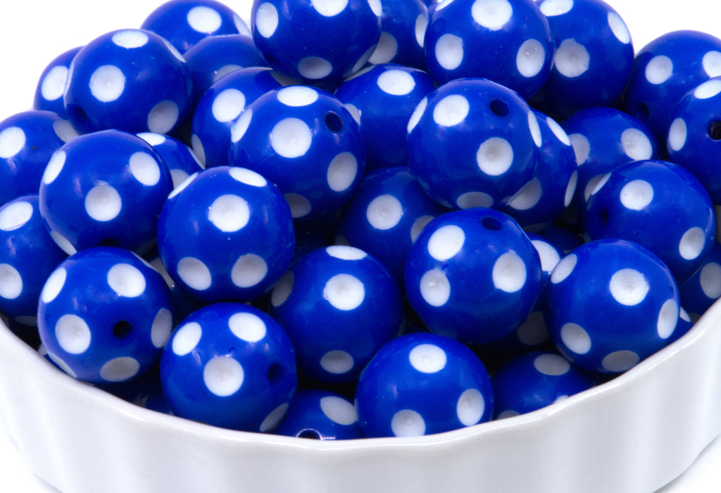 A close-up of blue round beads with white polka dots piled in a white bowl against a white background.