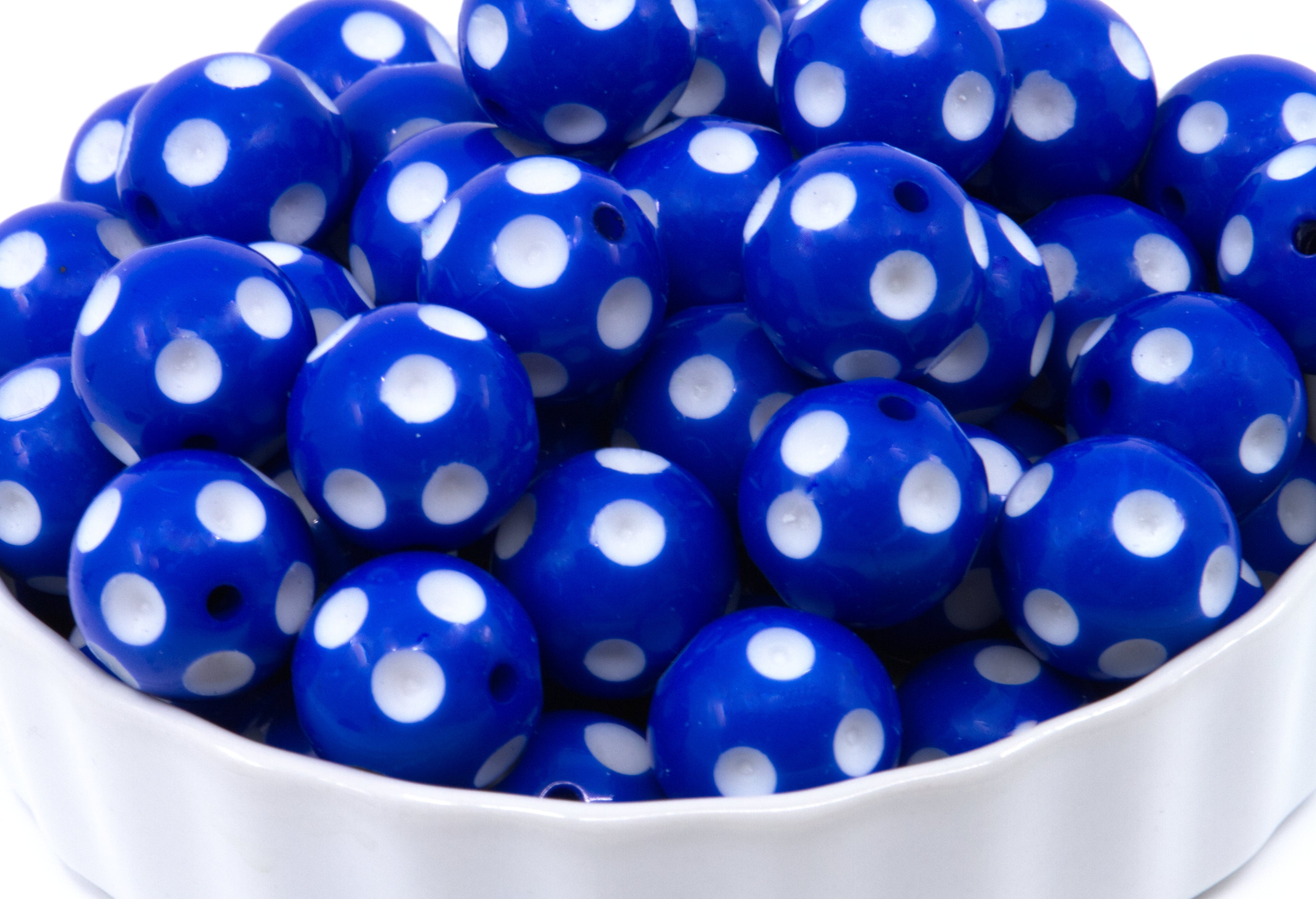 A close-up of blue round beads with white polka dots piled in a white bowl against a white background.