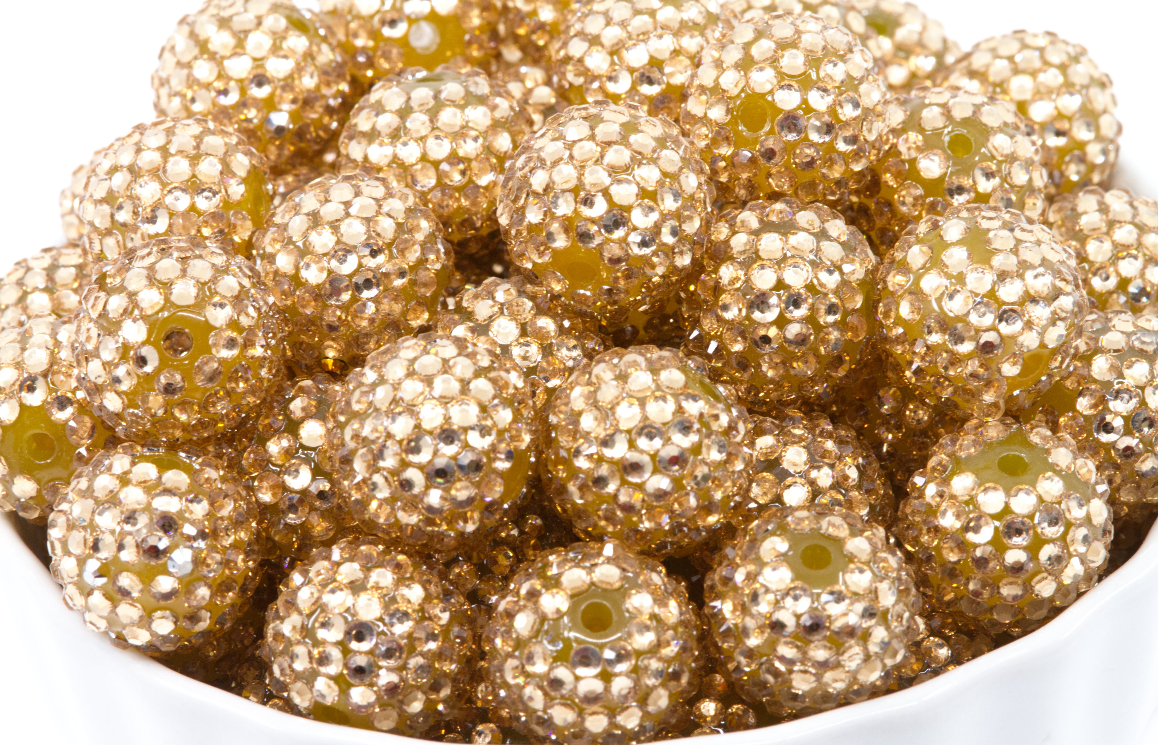 A close-up pile of round, champagne colored bubblegum beads covered in small reflective rhinestones, filling most of the frame against a white background.