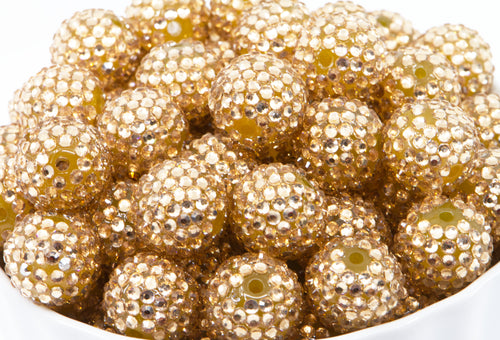 A close-up pile of round, champagne colored bubblegum beads covered in small reflective rhinestones, filling most of the frame against a white background.