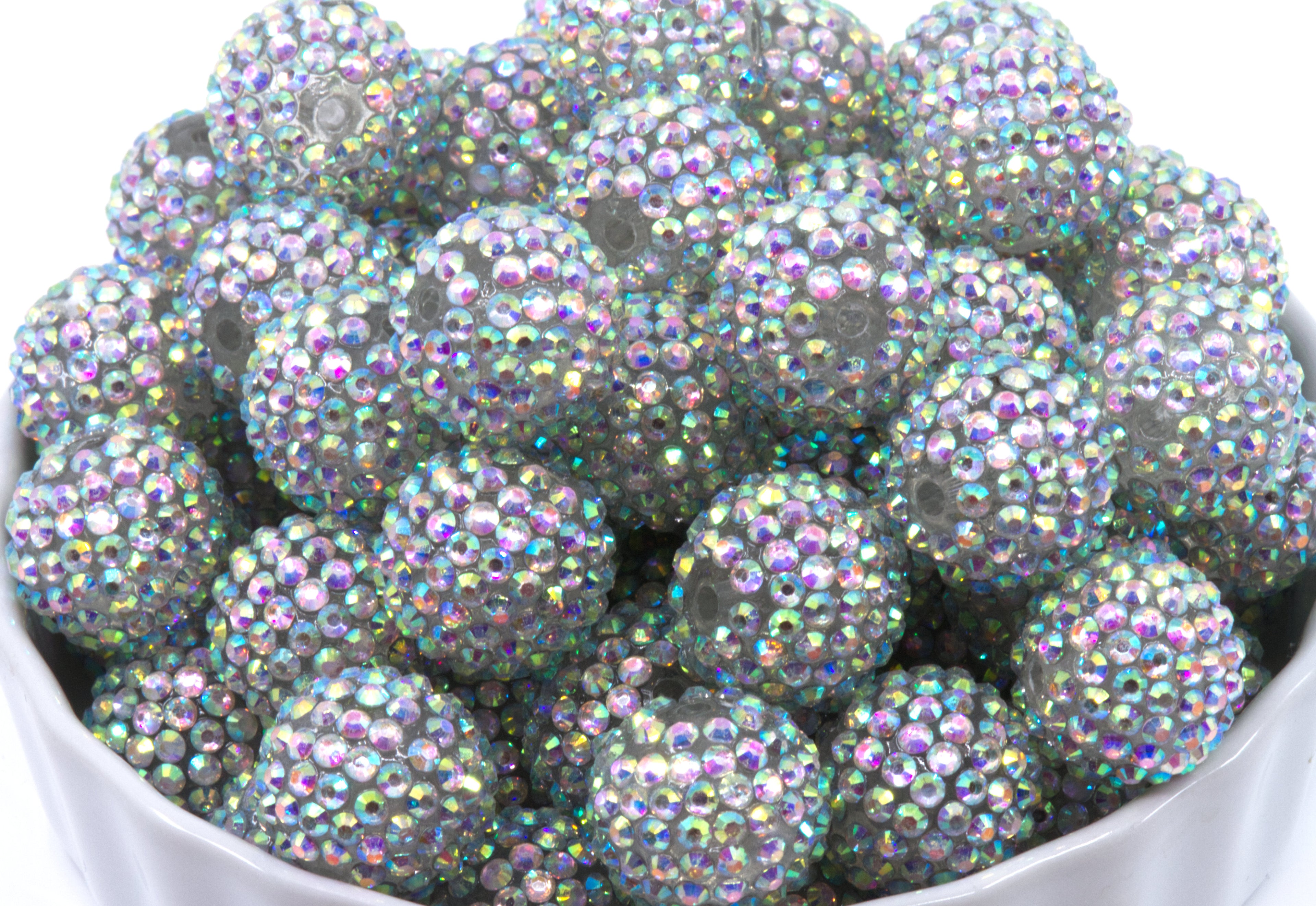 A pile of round, crystal AB rhinestone beads sitting in a white bowl against a white background.