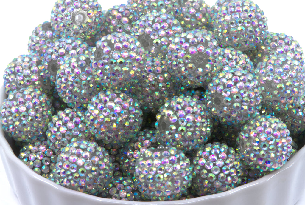 A pile of round, crystal AB rhinestone beads sitting in a white bowl against a white background.