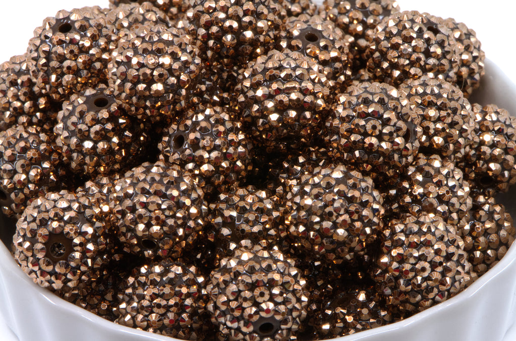 A close-up of a pile of rose gold rhinestone beads, piled in a white bowl against a white background.