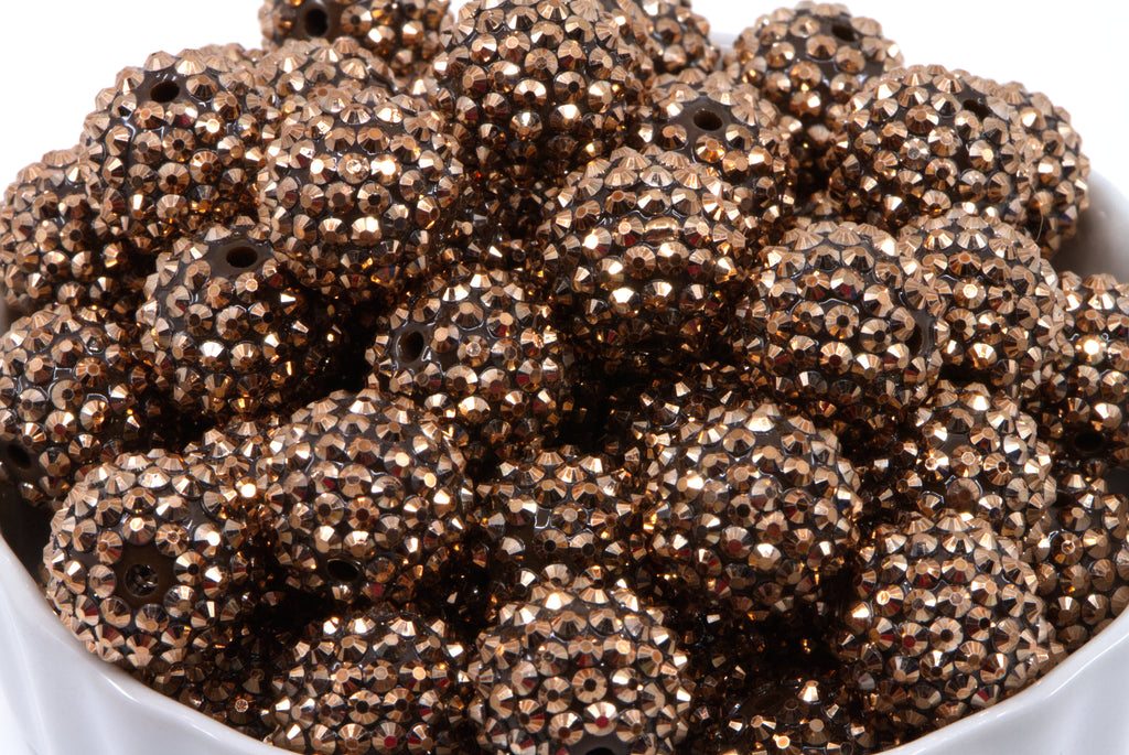 A close-up of a pile of rose gold rhinestone beads, piled in a white bowl against a white background.