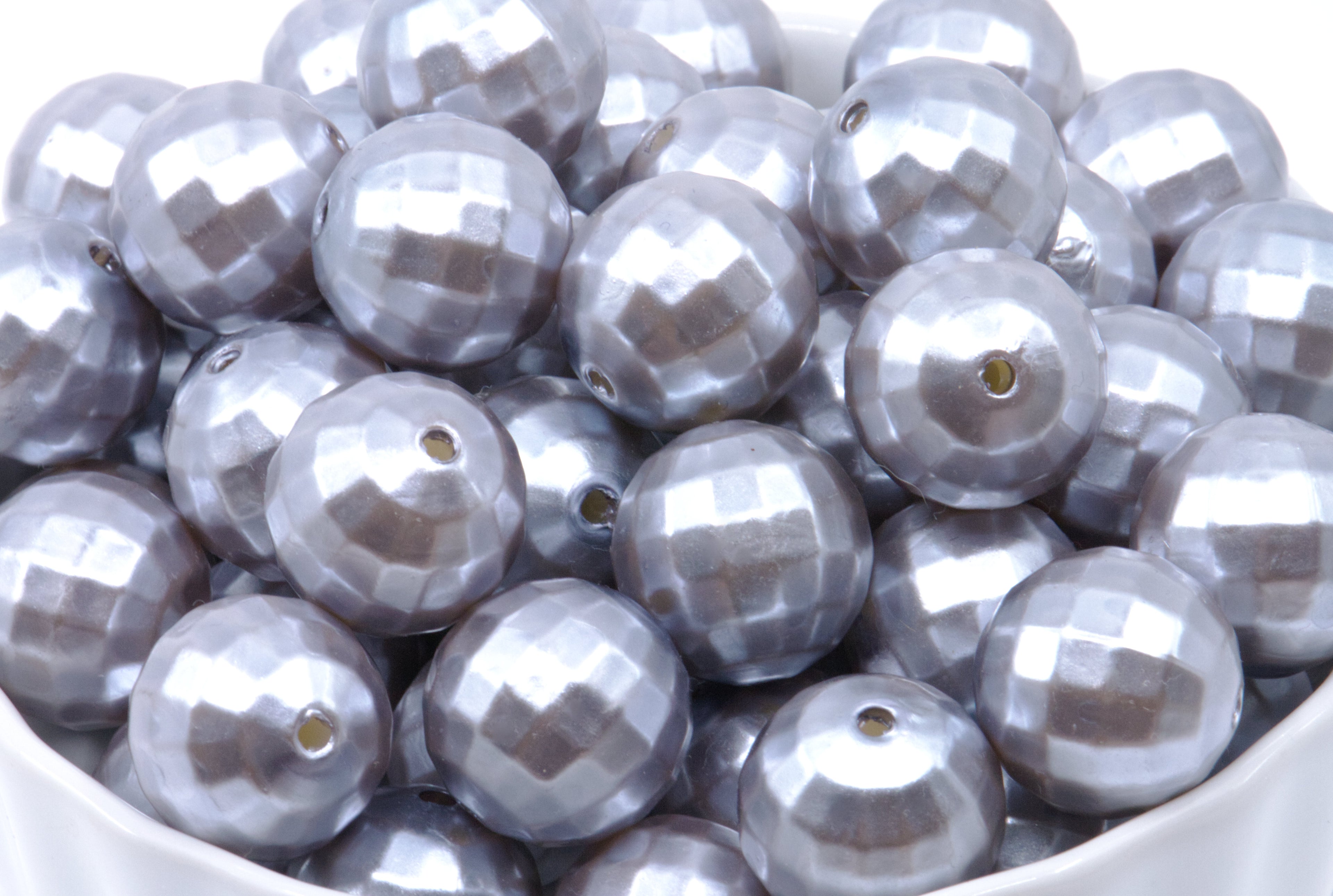 A close-up of shiny metallic silver faceted beads with a central hole, piled inside a white container against a white background.