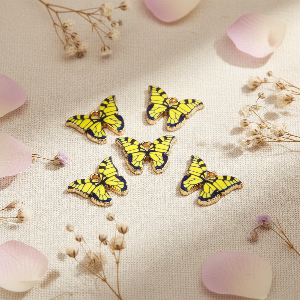 Five yellow and black butterfly-shaped jewelry charms with gold-tone edges, arranged on a textured cream fabric with scattered pink petals and dried flowers.