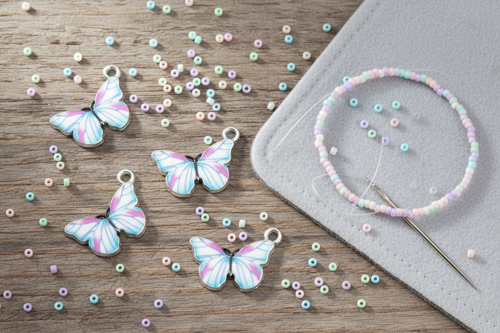Four butterfly charms with silver rings are arranged on a wooden surface alongside loose multicolored seed beads and a gray felt pad with a beaded bracelet and needle.