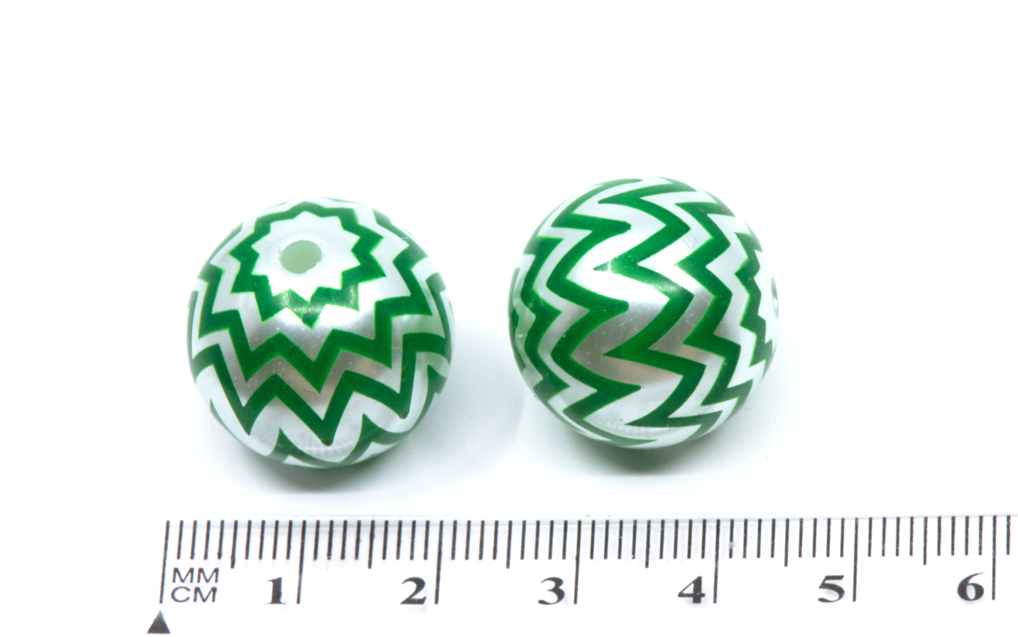 Two round bubblegum beads with a green and silver chevron pattern, shown next to a centimeter ruler for scale on a white background.