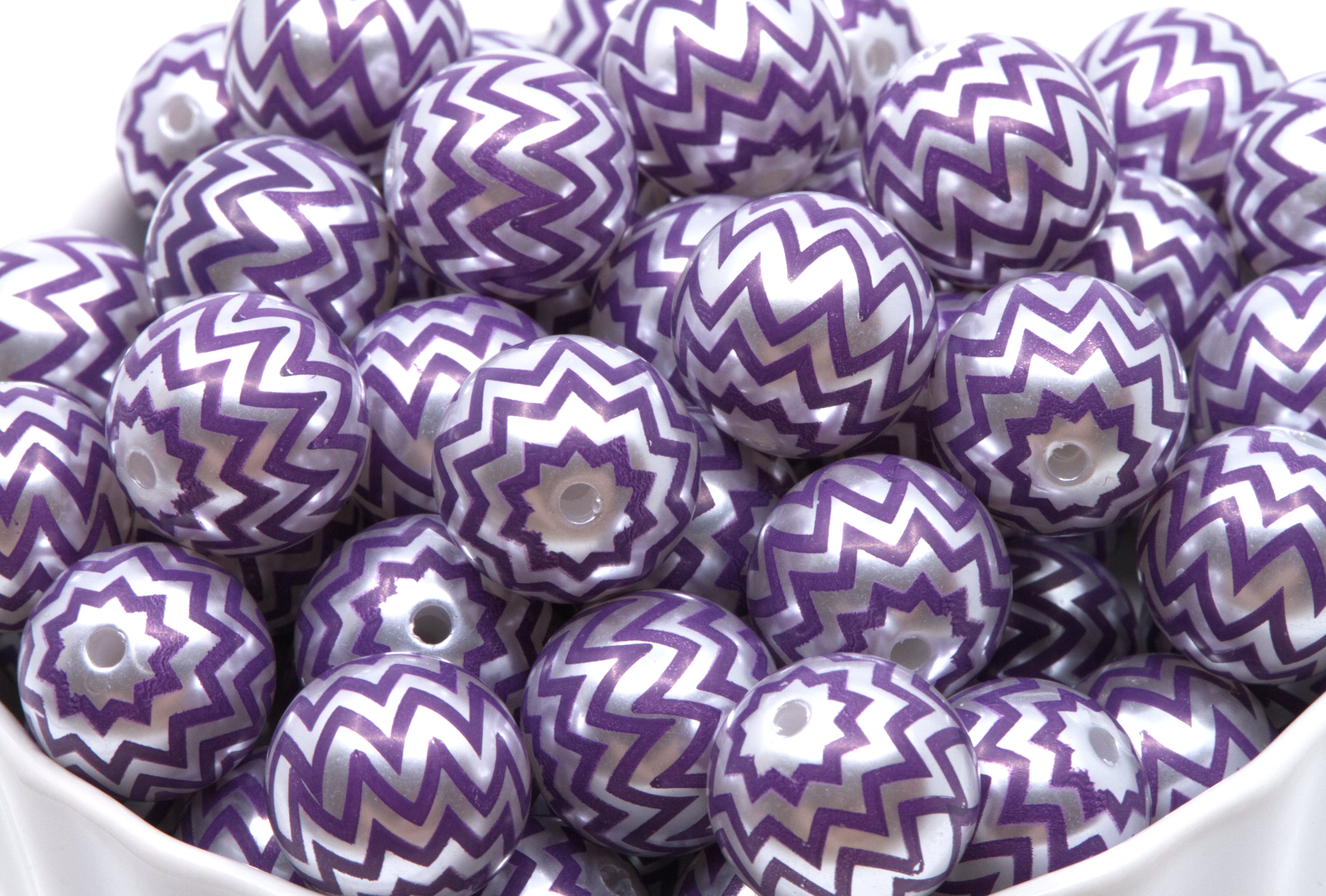 A pile of round craft beads with a purple and silver chevron pattern, shown in a white container.