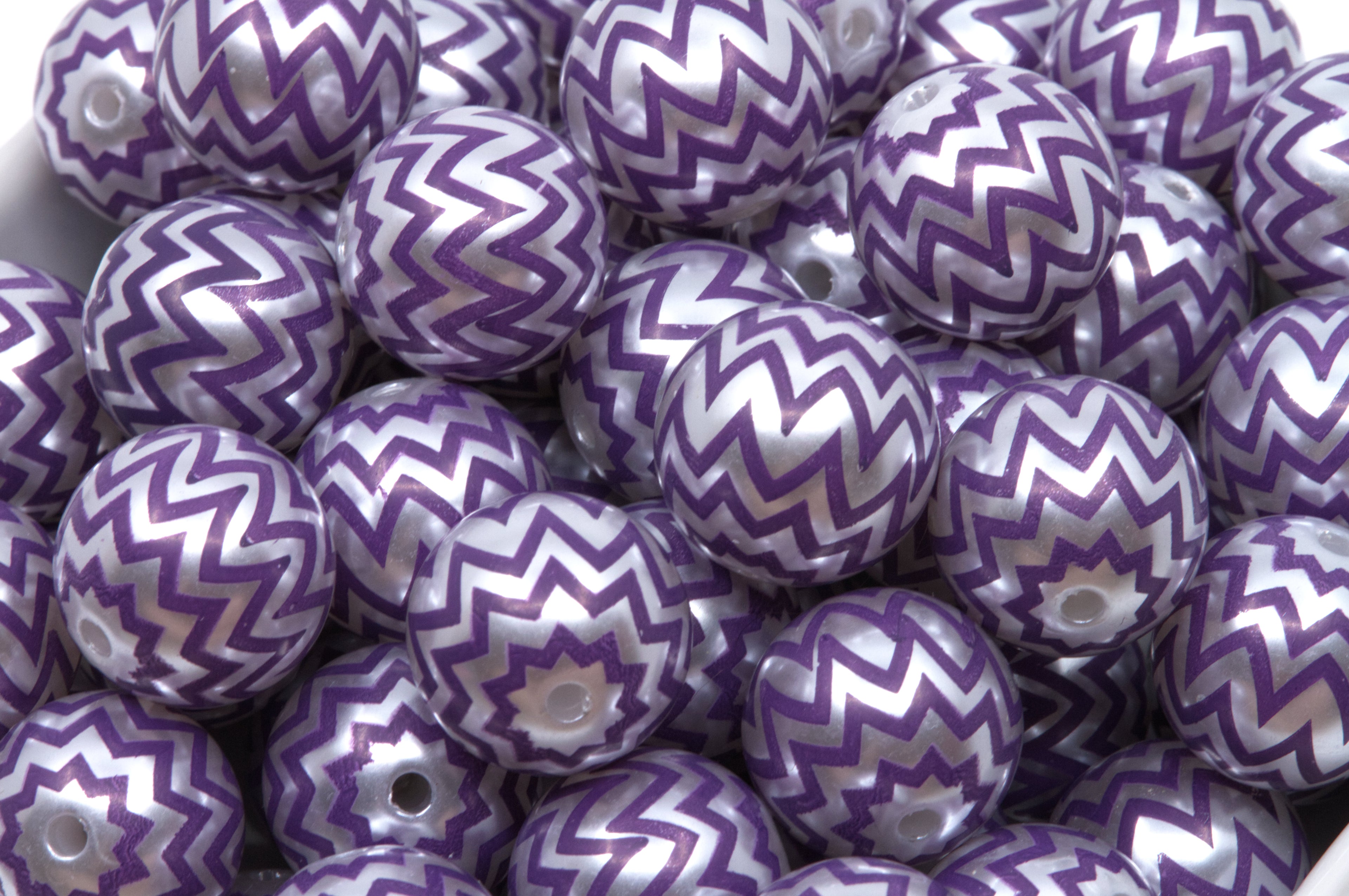 A pile of round craft beads with a purple and silver chevron pattern, shown in a white container.