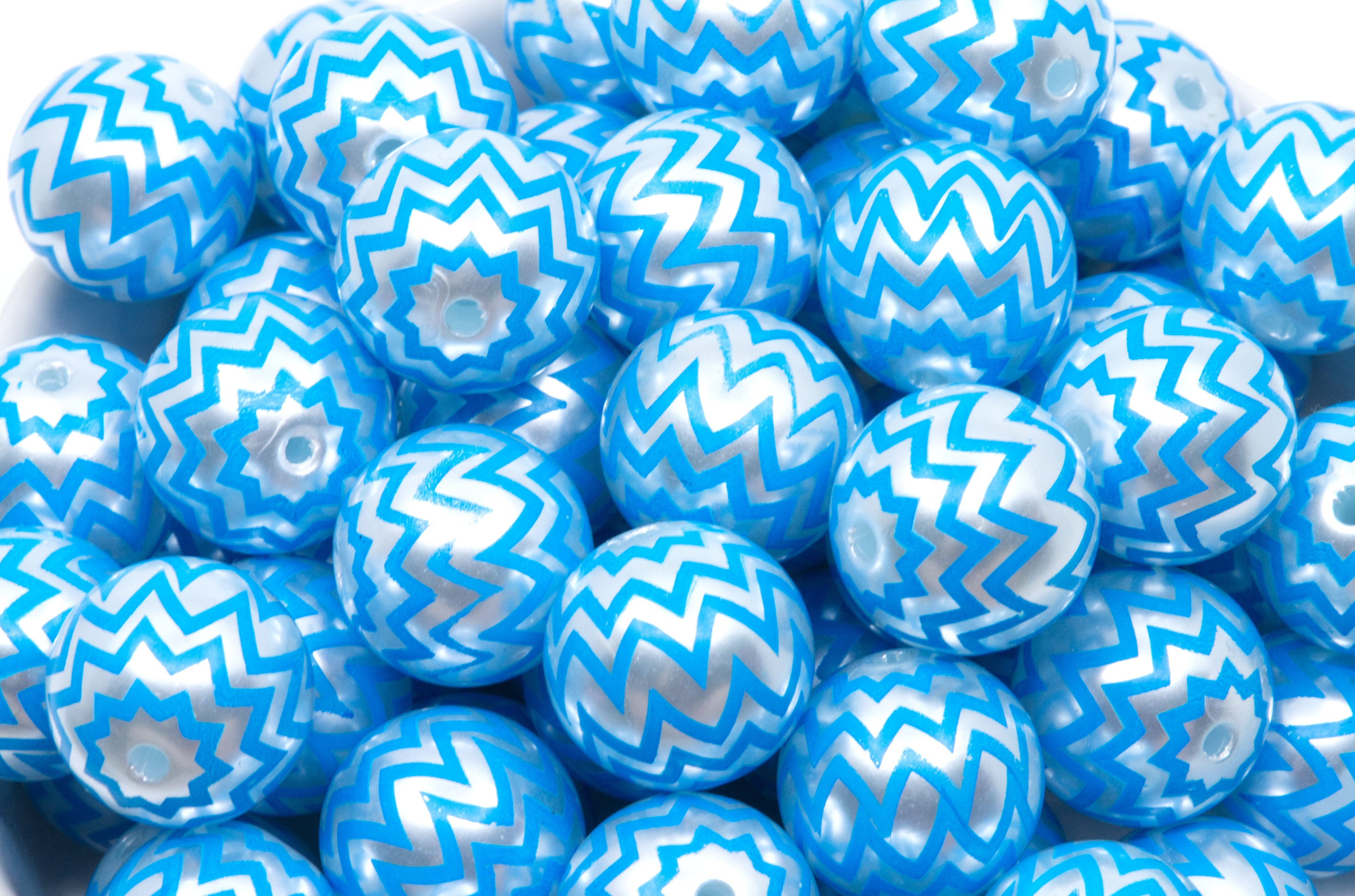 A close-up pile of  blue beads with a silver chevron zigzag pattern in a white bowl. 