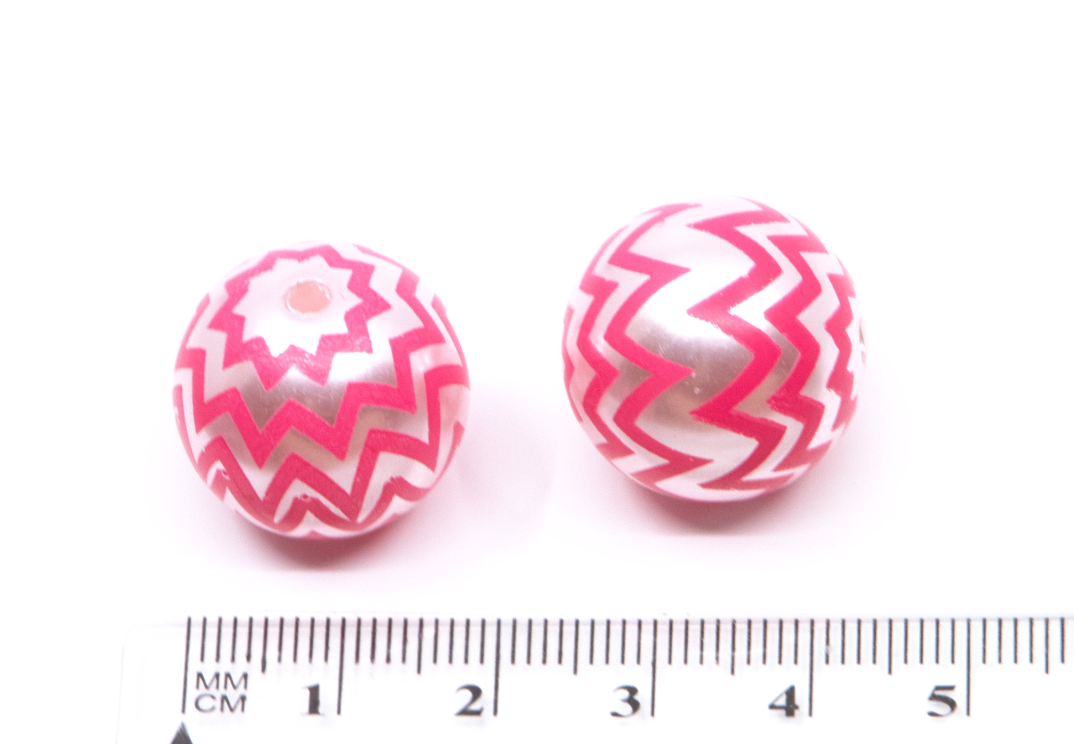 two round pink and silver chevron patterned beads next to a cm ruler for scale on a white background 