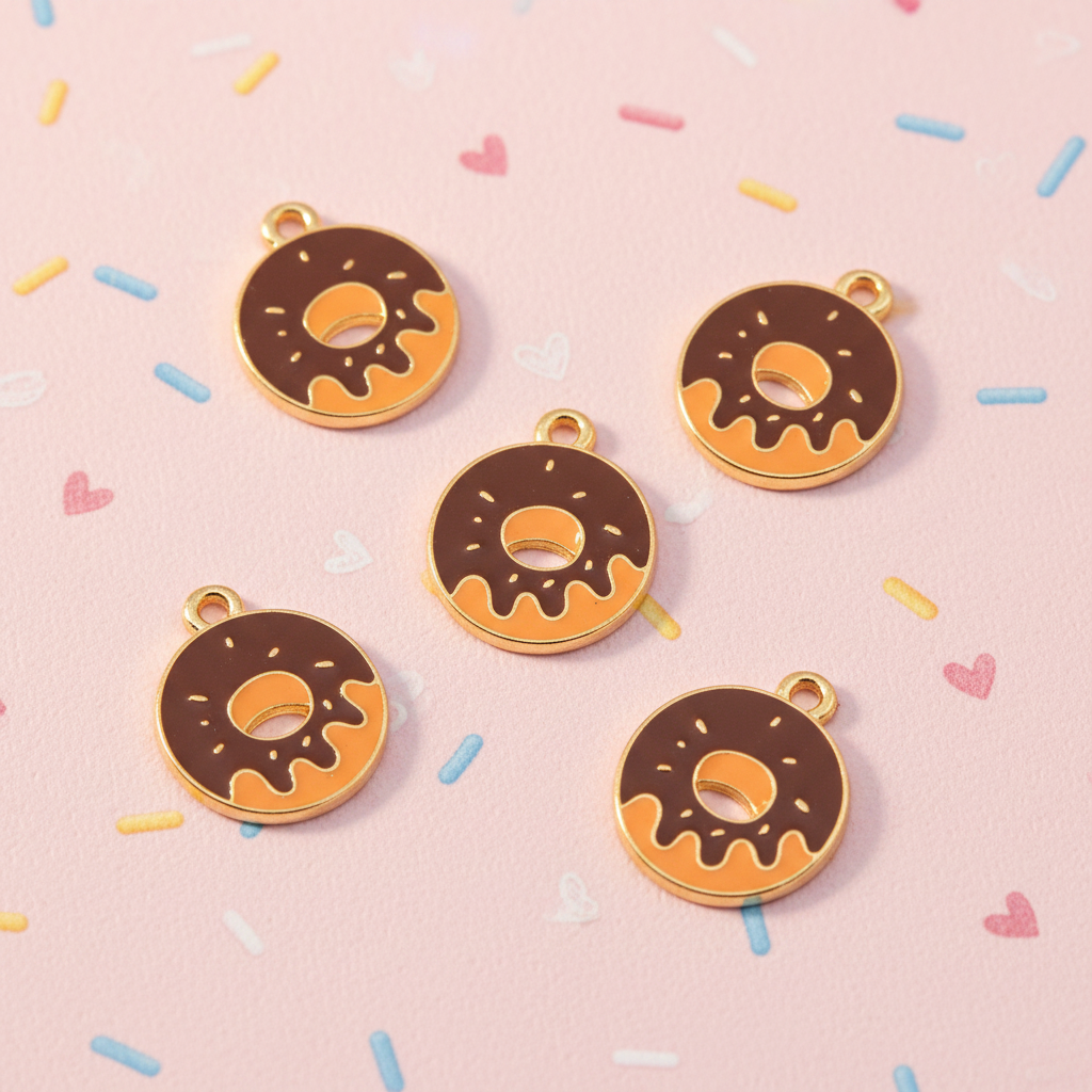 Five donut-shaped jewelry charms with gold-tone edges, brown chocolate frosting, arranged on a light pink background with scattered pastel sprinkles.