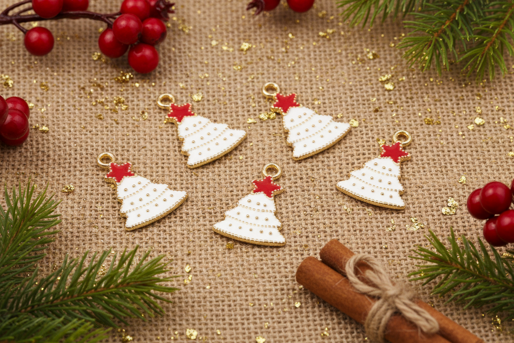 Five Christmas tree-shaped jewelry charms with gold-tone borders, white enamel, and red star tops, arranged on a burlap surface with pine branches, red berries, and cinnamon sticks.